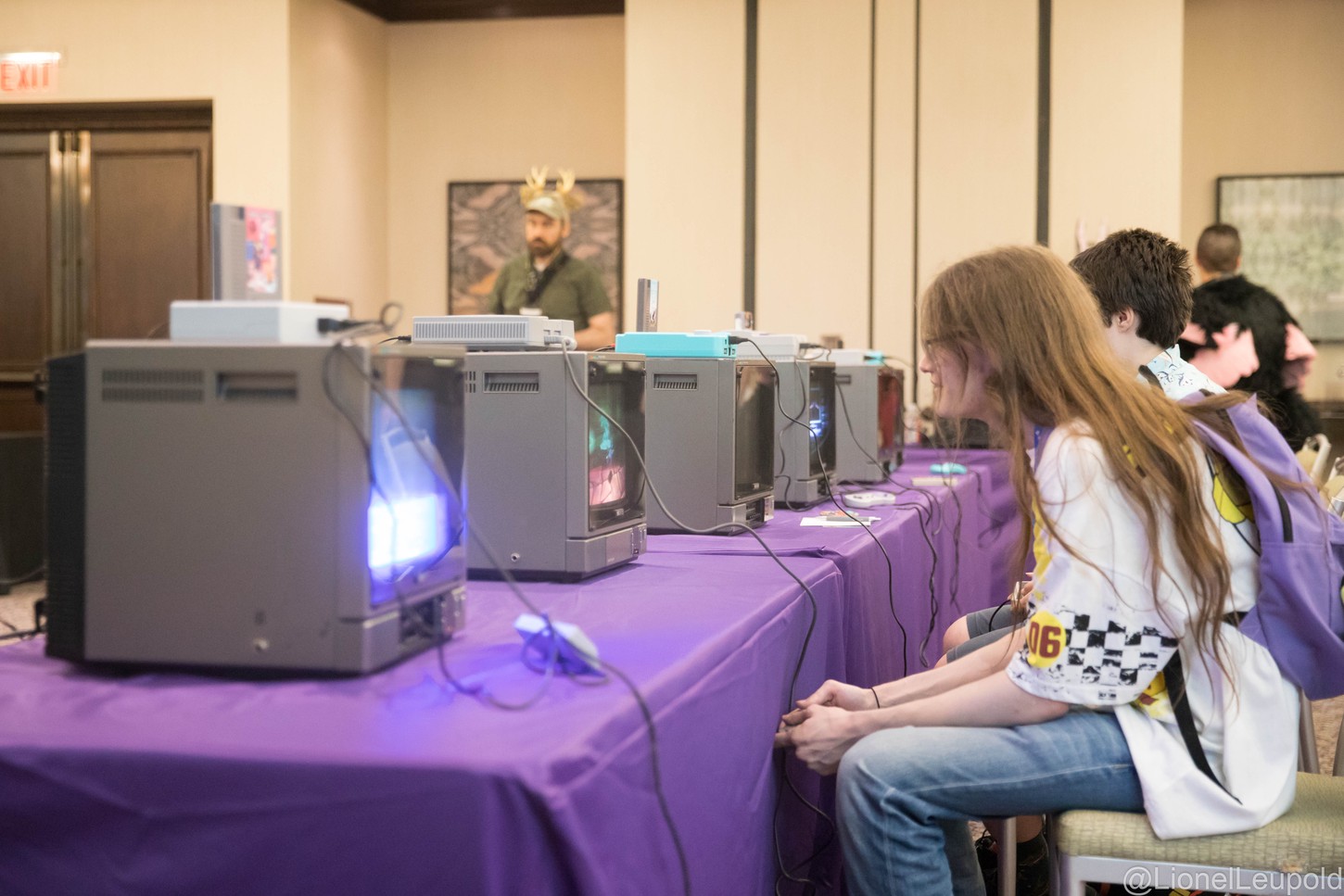 A photograph of a lineup of retro gaming consoles with CRT televisions set up in a room with people sitting in front of them playing the games.