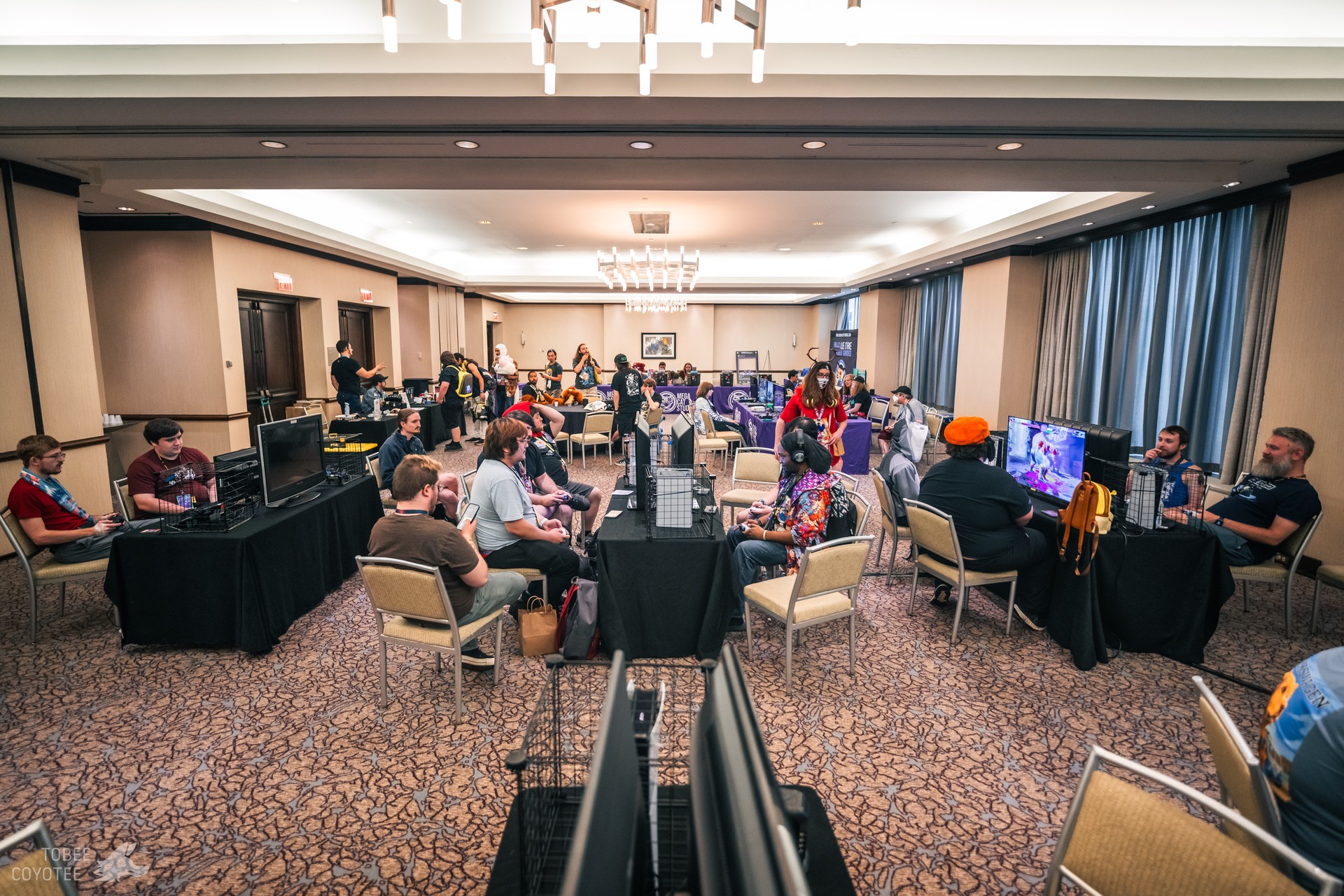 A photograph of a convention room. There are many tables set up with chairs surrounding them, each set up with televisions and gaming consoles. People are sitting at the tables, playing video games.